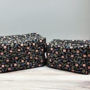 May include: Two black floral print zippered cosmetic bags. The bags feature a repeating pattern of pink, blue, and white flowers with green leaves. The bags are rectangular and have black zippers.