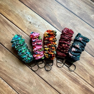 May include: Five fabric key fobs in various patterns and colors, including green with a floral design, pink with cherries, orange with stars, burgundy with white dots, and teal with a geometric pattern. Each has a metal clip and key ring.