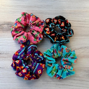 May include: Four colorful fabric scrunchies. One is pink with a cactus print, another is black with a geometric pattern, a third is purple with a similar geometric design, and the last is turquoise with a floral pattern. These hair accessories are a fun way to style your hair.
