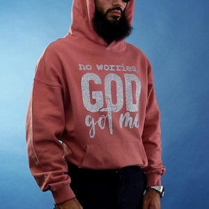 Può includere: Una felpa con cappuccio rosa polveroso con la scritta "no worries, GOD got me" in glitter argentati. La felpa ha una tasca frontale e un cappuccio. La persona indossa occhiali da sole neri e un orologio.