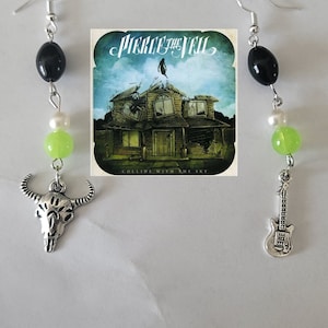 May include: A pair of earrings with black and green beads, a silver bull skull charm, and a silver guitar charm. The earrings are hanging from silver hooks. The album art for Pierce the Veil's "Collide with the Sky" is in the background.