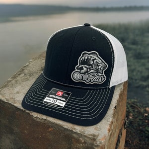 May include: Black and white trucker hat with a white mesh back. The hat features a patch with a bass fish and the text "Only Fins". The hat also has a red and white label with the text "Richardson 112 Trucker Adjustable".