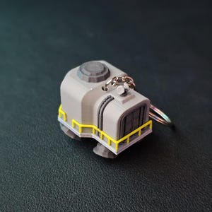 May include: A gray 3D printed keychain shaped like a small building with a yellow railing and a silver chain.