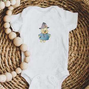 May include: White baby onesie with a cartoon image of Winnie the Pooh wearing a witch hat and a blue robe. The hat has pink and purple flowers on it.