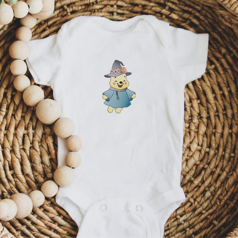 May include: White baby onesie with a cartoon image of Winnie the Pooh wearing a witch hat and a blue robe. The hat has pink and purple flowers on it.
