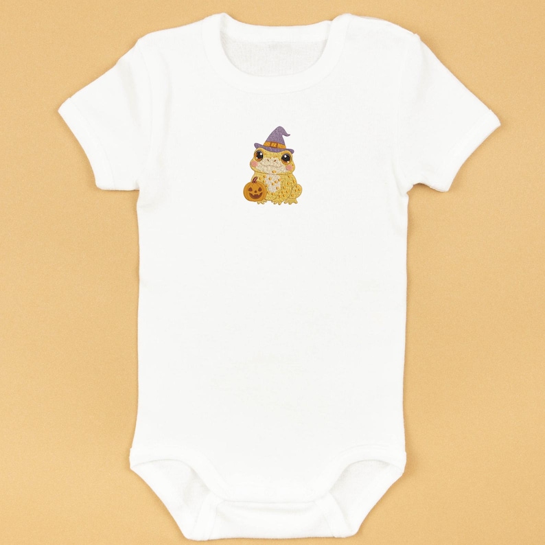 Puede incluir: Body de beb&eacute; blanco con una rana de dibujos animados que lleva un sombrero de bruja morado y sostiene una calabaza. La rana es amarilla y naranja, con ojos grandes. El body es de material suave, ideal para Halloween.