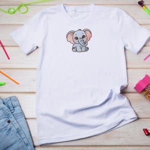Puede incluir: Camiseta blanca con un dise&ntilde;o de elefante de dibujos animados brillante. El elefante est&aacute; representado en azul y rosa, con orejas grandes y una trompa. La camiseta est&aacute; hecha de un material suave y es adecuada para ni&ntilde;os.