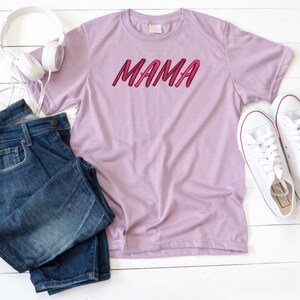 May include: A light purple t-shirt with the word "MAMA" printed in pink on the front. The shirt is laid flat on a white surface with a pair of blue jeans and white sneakers.