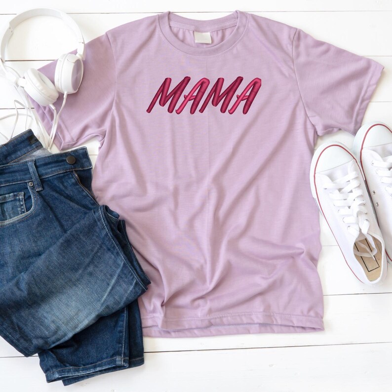 May include: A light purple t-shirt with the word "MAMA" printed in pink on the front. The shirt is laid flat on a white surface with a pair of blue jeans and white sneakers.