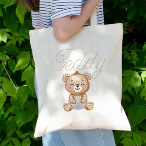 May include: A white canvas tote bag with a brown embroidered teddy bear holding a blue blanket. The bag has the name "Teddy" embroidered in brown cursive above the bear.