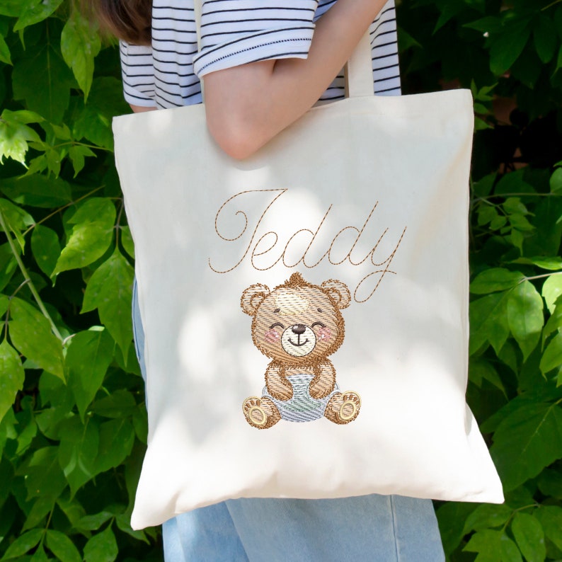 May include: A white canvas tote bag with a brown embroidered teddy bear holding a blue blanket. The bag has the name "Teddy" embroidered in brown cursive above the bear.