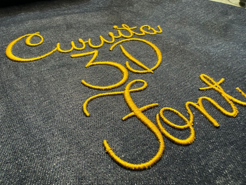 May include: A close-up of a dark blue fabric with the words "Curvita 3D Font" embroidered in yellow thread.