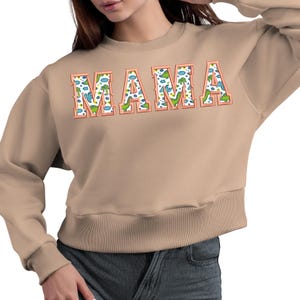 May include: A tan sweatshirt with a large graphic print of the word "MAMA" in a colourful dinosaur pattern.