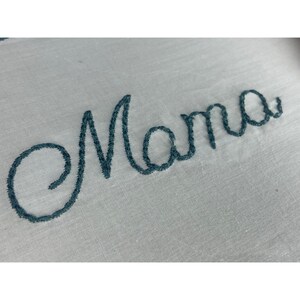 K&ouml;nnte beinhalten: Gestickter kursiv geschriebener Text "Mama" in blaugr&uuml;nem Garn auf wei&szlig;em Stoff.