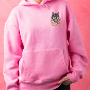 Puede incluir: Sudadera con capucha rosa con bolsillo delantero y capucha. La sudadera presenta un dise&ntilde;o bordado de la cabeza de un lobo rodeada de flores. El lobo es gris y negro, y las flores son rosas y amarillas.