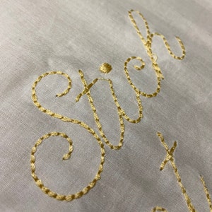 May include: Close-up of the word "stitch" embroidered in gold thread on a white fabric background.