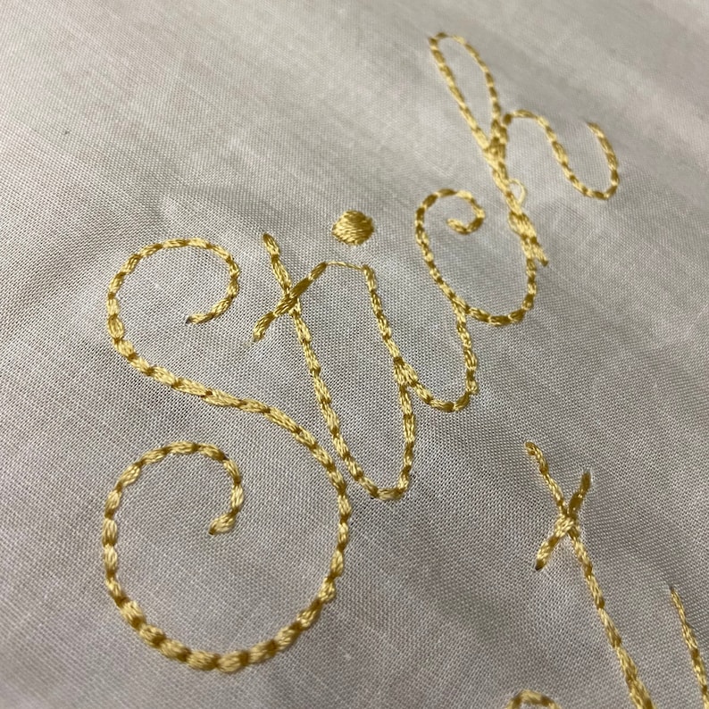 May include: Close-up of the word "stitch" embroidered in gold thread on a white fabric background.