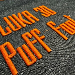 以下が含まれることがあります： 濃い青色の布地に、オレンジ色の刺繍で「Like a Puff of Fool」と書いてあります。