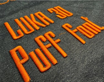 May include: Orange embroidered text on a dark blue fabric background that reads "Like a Puff of Fool".