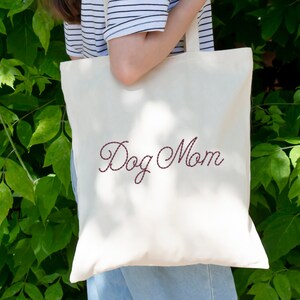 K&ouml;nnte beinhalten: Eine wei&szlig;e Canvas-Tragetasche mit dem Schriftzug "Dog Mom", der in maroonfarbener Farbe gestickt ist.