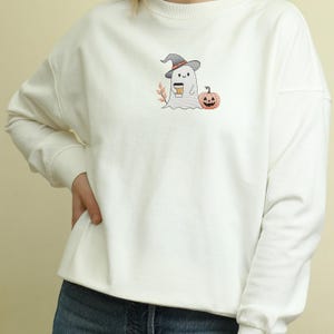 May include: White sweatshirt with a cute embroidered ghost wearing a witch hat and holding a cup of coffee. The ghost is standing next to a small orange pumpkin.