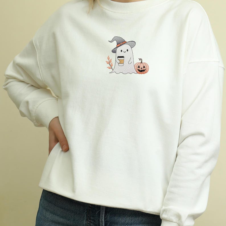 May include: White sweatshirt with a cute embroidered ghost wearing a witch hat and holding a cup of coffee. The ghost is standing next to a small orange pumpkin.