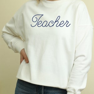 K&ouml;nnte beinhalten: Wei&szlig;er Pullover mit dem Wort "Teacher" in blauer Stickerei.