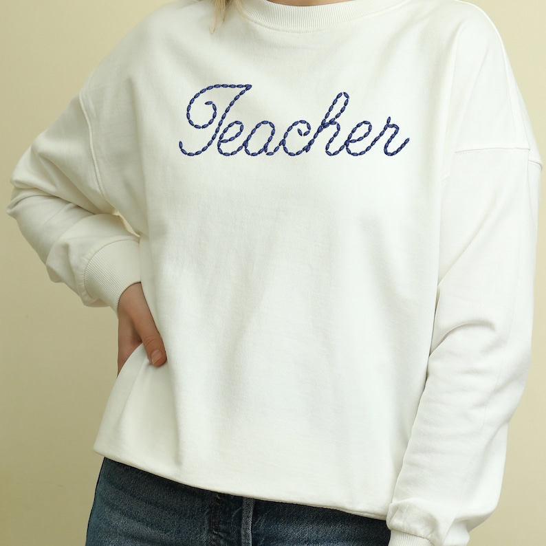 K&ouml;nnte beinhalten: Wei&szlig;er Pullover mit dem Wort "Teacher" in blauer Stickerei.