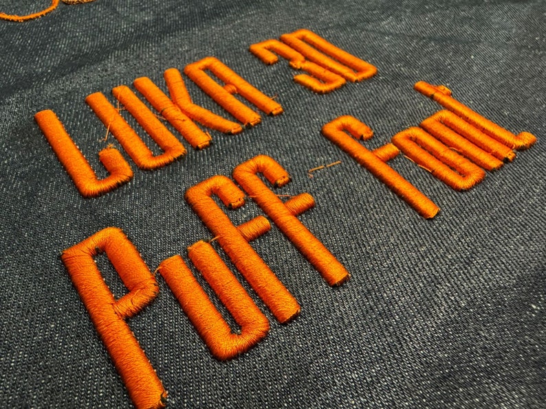 May include: Orange embroidered text on a dark blue fabric background that reads "Like a 3D Puff Font".