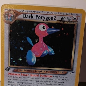 May include: A Dark Porygon2 Pokemon card from the Pokemon Trading Card Game. The card is yellow with a holographic image of the Pokemon. The card has a blue and pink Pokemon with a long beak. The card text reads "Dark Porygon2 60 HP Stage 1 Pokemon".