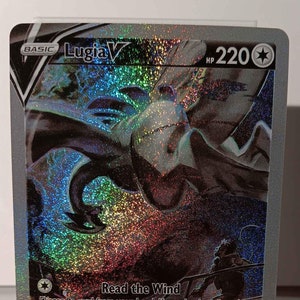 May include: A holographic Pokemon trading card featuring Lugia V, a blue and white legendary Pokemon with a wingspan. The card has a silver background and features the text "Lugia V" and "HP 220". The card also has the text "Read the Wind" and "Aero Dive" with attack descriptions. The card has a V rule that states "When your Pokemon V is Knocked Out, your opponent takes 2 Prize cards."