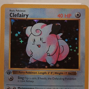 May include: A Clefairy Pokemon card from the first edition of the Pokemon Trading Card Game. The card is holographic and features a pink Clefairy with a pink bow. The card has a white background with a blue border. The card text reads "Basic Pokemon Clefairy 40 HP Fairy Pokemon. Length: 2' 0", Weight: 17 lbs. Sing Flip a coin. If heads, the Defending Pokemon is now Asleep. Metronome Choose 1 of the Defending Pokemon's attacks, Metronome copies that attack except for its Energy costs and anything else required in order to use that attack, such as discarding Energy cards. (No matter what type the Defending Pokemon is, Clefairy's type is still Colorless.) Weakness Resistance -30 Retreat Cost Its magical and cute appeal has many admirers. It is rare and found only in certain areas. LV. 14 #35 Illus. Ken Sugimori 1995, 96, 98, 99 Nintendo, Creatures, GAMEFREAK 1999 Wizards. 5/102"