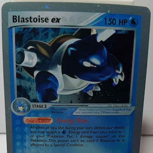 May include: A Pokémon trading card featuring Blastoise ex with 150 HP. The card has a blue and silver design with a Blastoise illustration. Text includes "Hyper Whirlpool" and game instructions. The card is on a white stand.