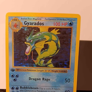 May include: A holographic Gyarados Pokémon card. The card features a green and yellow serpentine creature with a red tongue. The card has a blue background with text that reads "Gyarados" and "100 HP". The card is on a clear display stand.