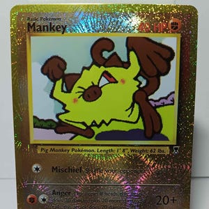 May include: A holographic trading card featuring the Pokémon Mankey. The card has a gold foil background and displays a cartoon image of Mankey, a yellow and brown monkey-like creature. The card text includes the Pokémon's name, stats, and attack details.