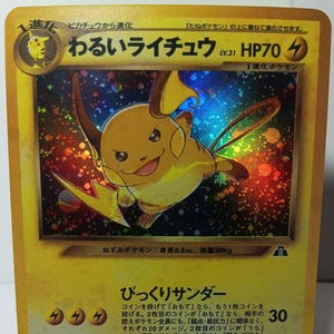 May include: A Dark Raichu trading card featuring a holographic illustration of the yellow electric-type creature. The card displays the name in Japanese, HP70, and lightning energy symbols. The background of the illustration has a colorful, sparkling foil effect.