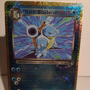 May include: A holographic Dark Blastoise Pokémon card. The card features a blue Blastoise character with a cannon on its back. The card has text that reads "Dark Blastoise 70 HP" and "Shellfish Pokémon. Length: 5' 3", Weight: 189 lbs."