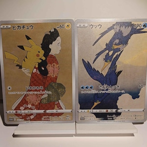 May include: Two Pokémon trading cards. One features Pikachu with a woman in a red kimono, the other depicts blue birds. Both cards have Japanese text and are displayed on white stands.