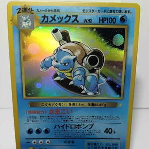 May include: A holographic Pokémon trading card featuring Blastoise, a blue turtle-like creature. The card has Japanese text and shows the character's HP, level, and attack details. The card is displayed in a white stand.