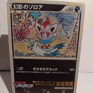 May include: A Pokémon trading card featuring a colorful illustration of a Zorua character with other cartoon figures. The card has Japanese text and a drawing of the Eiffel Tower. The card is displayed on a white stand.