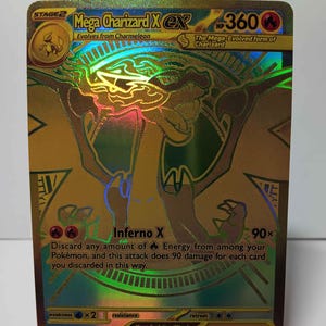 May include: A gold holographic Mega Charizard X EX Pokémon trading card. The card features a detailed illustration of Charizard in a dynamic pose, with the text "Mega Charizard X EX" and "Inferno X" visible. The card is displayed on a white stand.