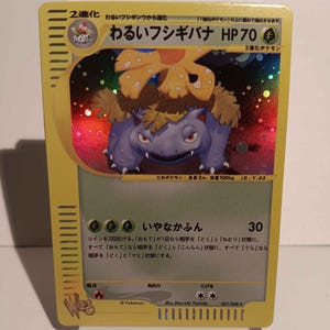 May include: A Pokémon trading card featuring a purple and green creature with a large flower on its back. The card has Japanese text, the HP is 70, and the card is labeled as a 2-stage evolution. The card is on a white stand.