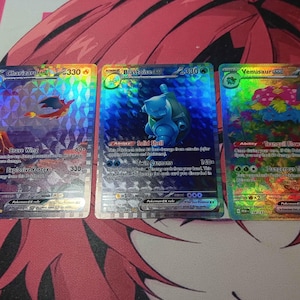 May include: Three holographic Pokémon trading cards featuring Charizard, Blastoise, and Venusaur. The Charizard card shows a fiery dragon, the Blastoise card features a blue turtle, and the Venusaur card displays a plant-like creature.