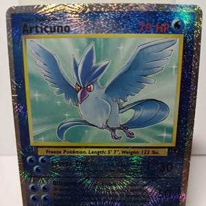 May include: A holographic Articuno Pokémon trading card. The card features a blue bird with outstretched wings, a red beak, and a blue crest. The card's text includes "Articuno", "Freeze Pokémon", and "Length: 5' 7", Weight: 122 lbs."