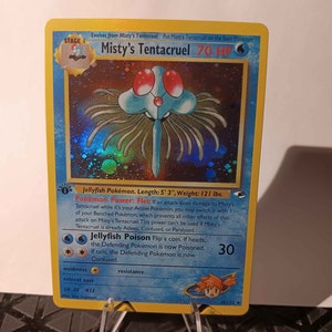 May include: A holographic Misty's Tentacruel Pokémon card. The card features a blue jellyfish-like creature with red eyes and a light blue body. The card has text describing the creature's abilities and stats, including a length of 5' 3" and a weight of 121 lbs.