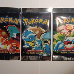 May include: Three sealed Pokémon trading card packs. Each pack features a different Pokémon character: Charizard, Blastoise, and Venusaur. The packs are dark blue with the Pokémon logo and text that reads "11 Tradable Game Cards."
