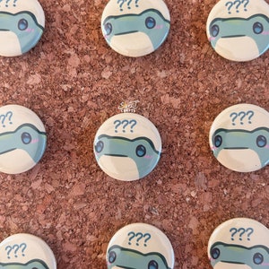 Könnte beinhalten: Neun runde Knöpfe mit einem Comic-Froschgesicht auf jedem. Der Frosch hat einen hellblauen Körper, weiße Augen und rosa Wangen. Die Knöpfe sind beige mit dem Text "???" in Schwarz über dem Frosch.