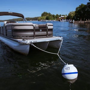 May include: A white pontoon boat with a brown and gray top, docked at a pier. The boat has a white rope attached to a white buoy with a blue stripe.