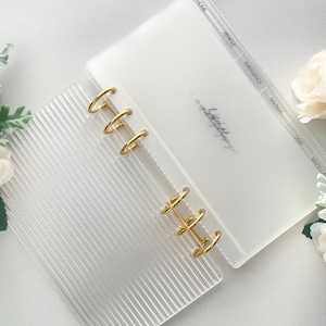 May include: A clear, open binder with gold rings and labeled dividers. The binder has a ribbed cover and includes sections for Home, Vehicle, Emergency, Savings, Debt, and Miscellaneous. The text "a homebody" is written on the inside cover.