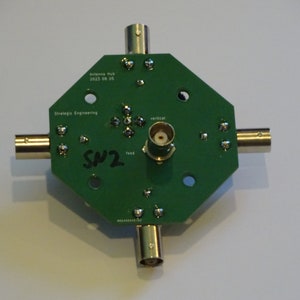 May include: A green circuit board with four F-type connectors and a center connector labeled "vertical". The board is labeled "Antenna Hub 2023 08 05" and "Strategic Engineering". The board is labeled "SW2" in black marker.